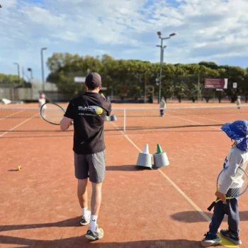Coach and a young player in a sun hat walking onto a clay court, seen from behind