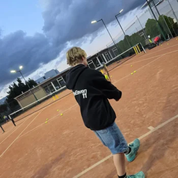 Junior player, seen from behind, on a clay court under an evening sky