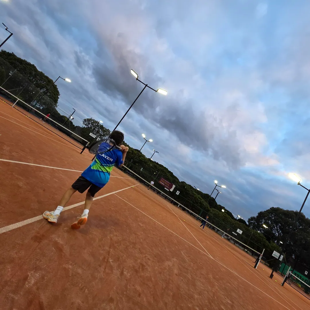Player on a clay court at dusk, seen from a distance
