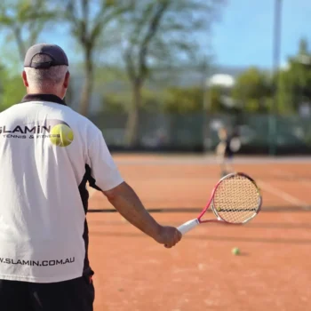 Slamin Tennis coach, seen from behind, feeding balls during a lesson