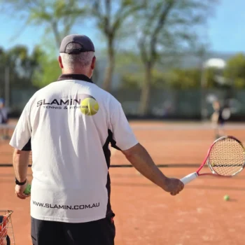 Slamin Tennis coach, seen from behind, feeding balls during a lesson on a clay court