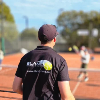 Slamin Tennis coach, seen from behind, watching a junior lesson on a clay court